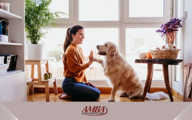 Eine Frau in einem gemütlichen Raum, die vor einem golden Retriever sitzt und ihm die Pfote gibt. Im Hintergrund sind Pflanzen und ein Tisch mit Dekorationen zu sehen.