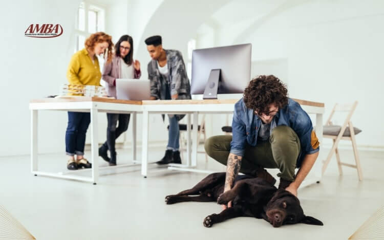 Eine Büro-Szene mit vier Personen, die an einem Tisch arbeiten, während ein Mann mit einem braunen Hund auf dem Boden spielt.