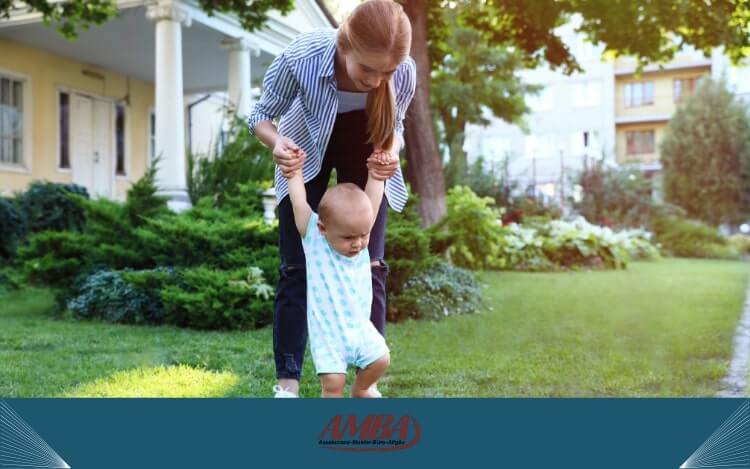 Eine Au Pair hält ein Kleinkind an den Händen, während es im Garten läuft. Die Umgebung ist grün und gut gepflegt, mit Bäumen und Sträuchern.