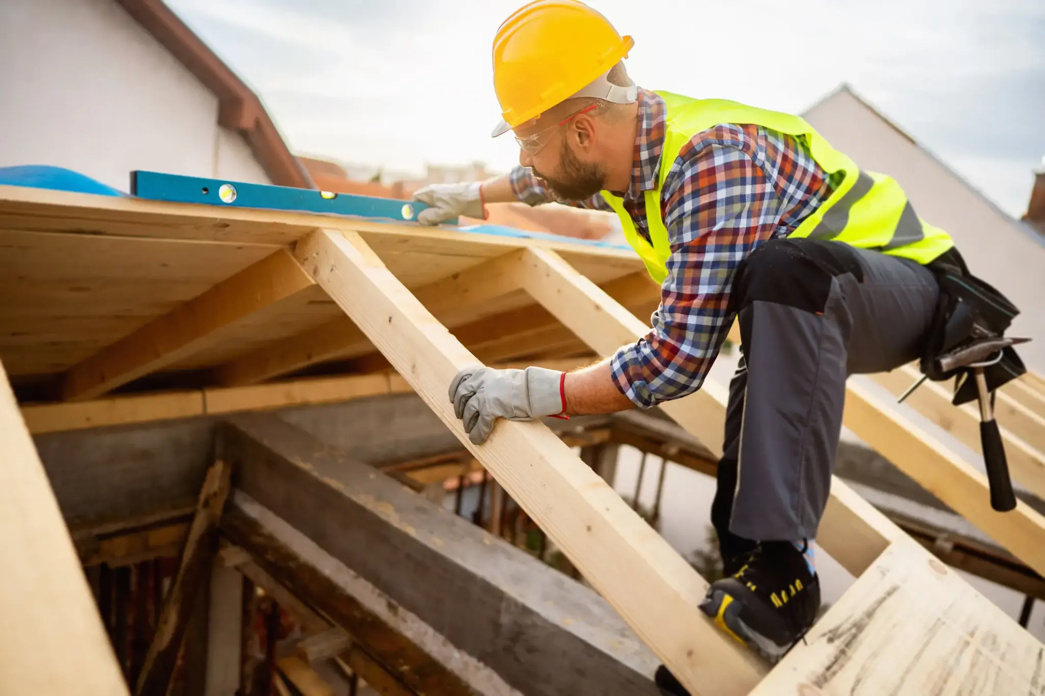 Dachdecker misst und richtet Holzbalken auf einem Neubau aus, mit Schutzkleidung, Helm und Wasserwaage bei sonnigem Wetter