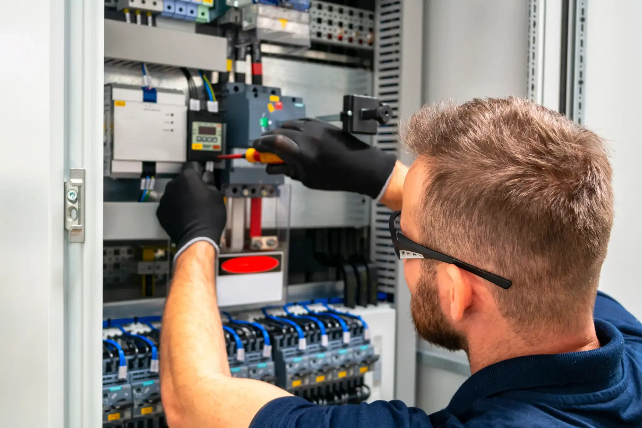 Elektriker bei der Arbeit an einem offenen Schaltschrank, mit Schraubendreher und Schutzhandschuhen