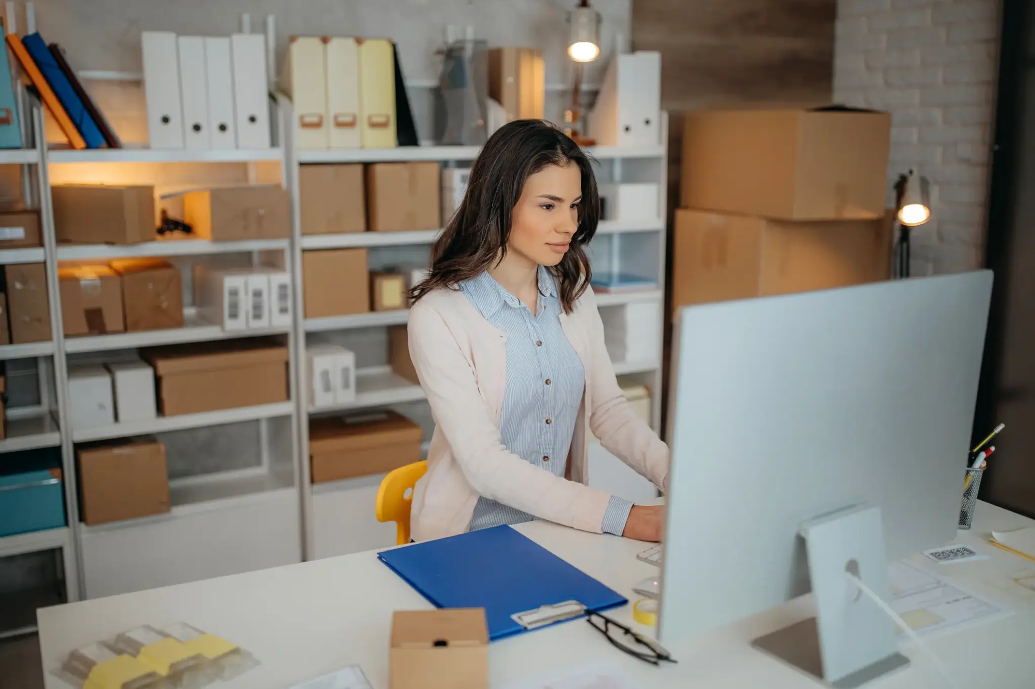 Junge Frau arbeitet konzentriert am Schreibtisch im Lager und organisiert ihr Kleingewerbe am Computer
