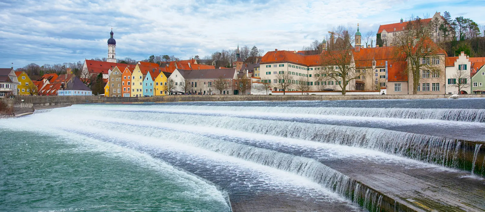 Landsberg | Wellenförmiges Lechwehr in Landsberg am Lech mit bunten Häusern und der historischen Altstadt im Hintergrund bei Tageslicht