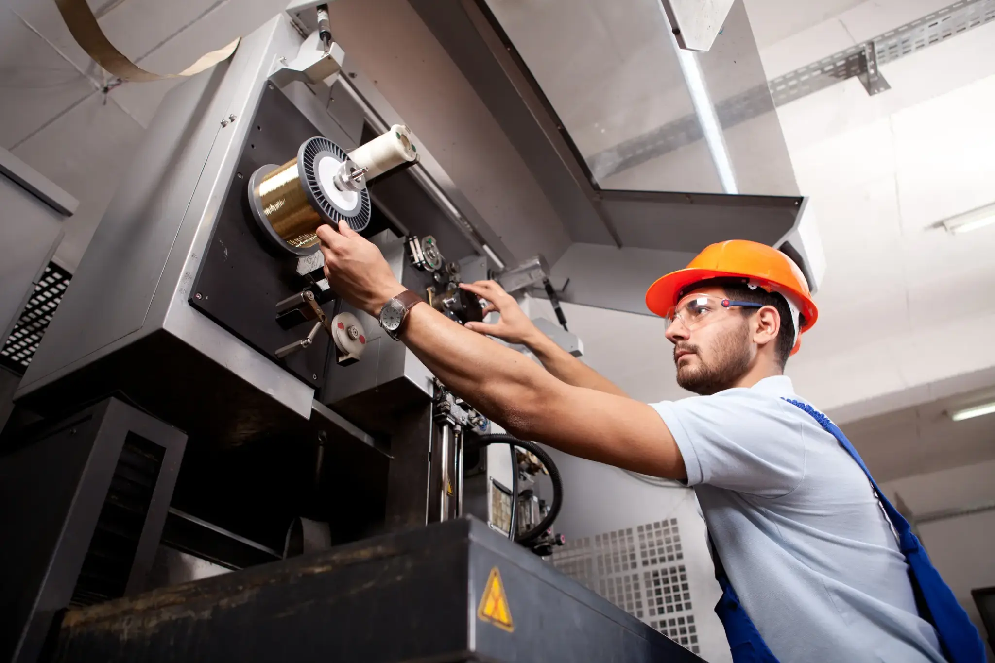Techniker mit Schutzhelm prüft in der Industriehalle eine laufende Produktionsmaschine auf Funktion und Sicherheit