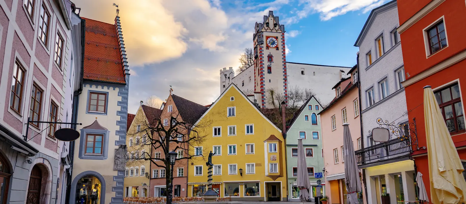 Bunte Häuserfassaden in der Altstadt von Füssen mit Blick auf das Hohe Schloss und den Uhrturm bei leicht bewölktem Himmel