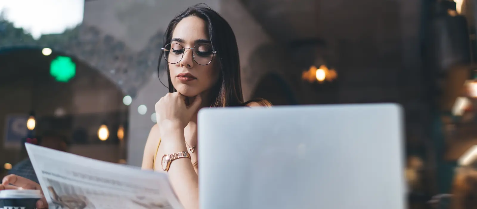 interview | Junge Frau mit Brille liest aufmerksam eine Zeitung in einem Café, vor ihr steht ein Laptop, im Hintergrund warme Lichtquellen