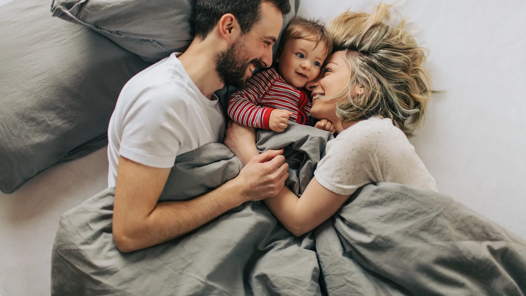 Junge Familie liegt gemeinsam im Bett und lacht, Mutter, Vater und Baby blicken sich liebevoll an