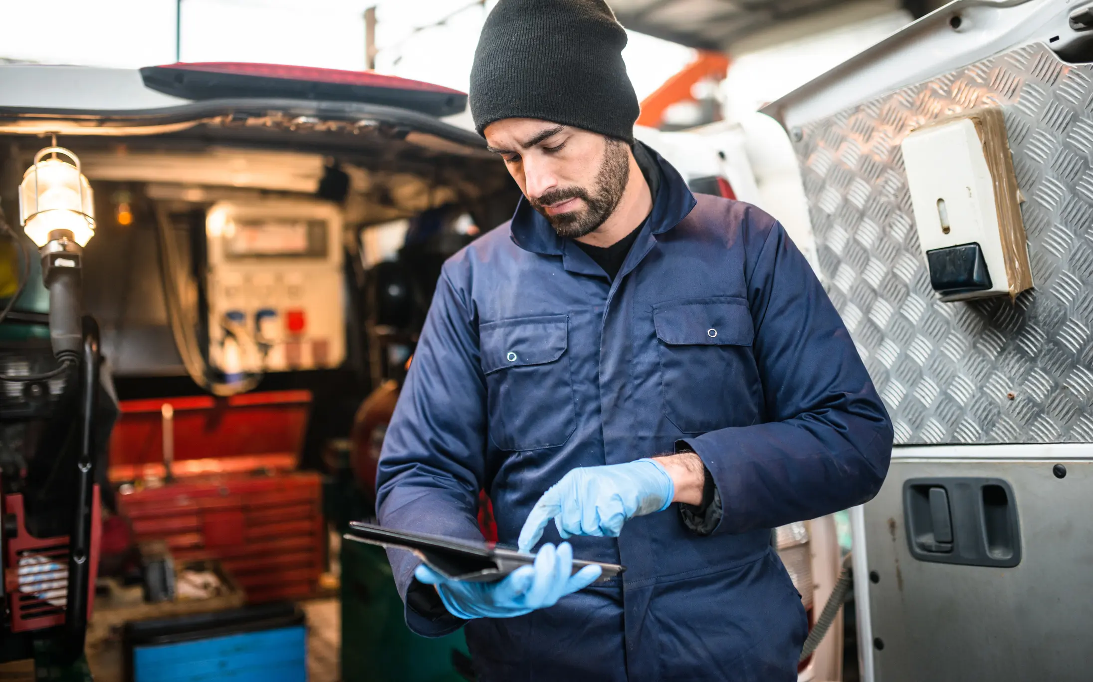Handwerker im blauen Overall bedient ein Tablet vor offenem Werkstattfahrzeug – Symbol für mobilen Werkverkehr und berufliche Absicherung