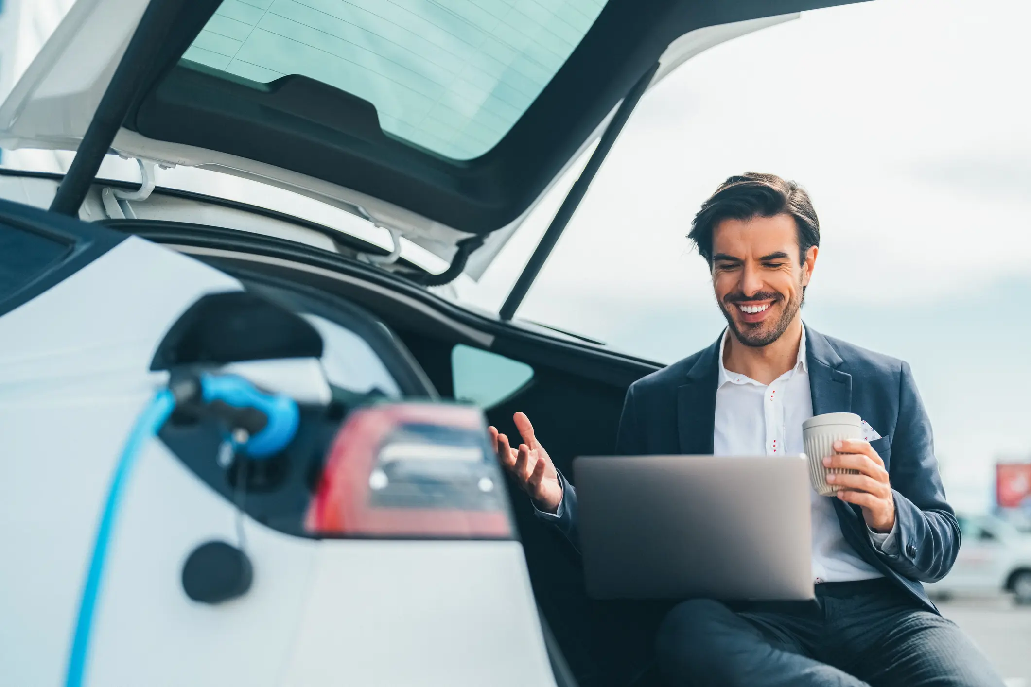Mann arbeitet am Laptop neben Elektro-Firmenwagen an Ladestation und hält eine Kaffeetasse in der Hand