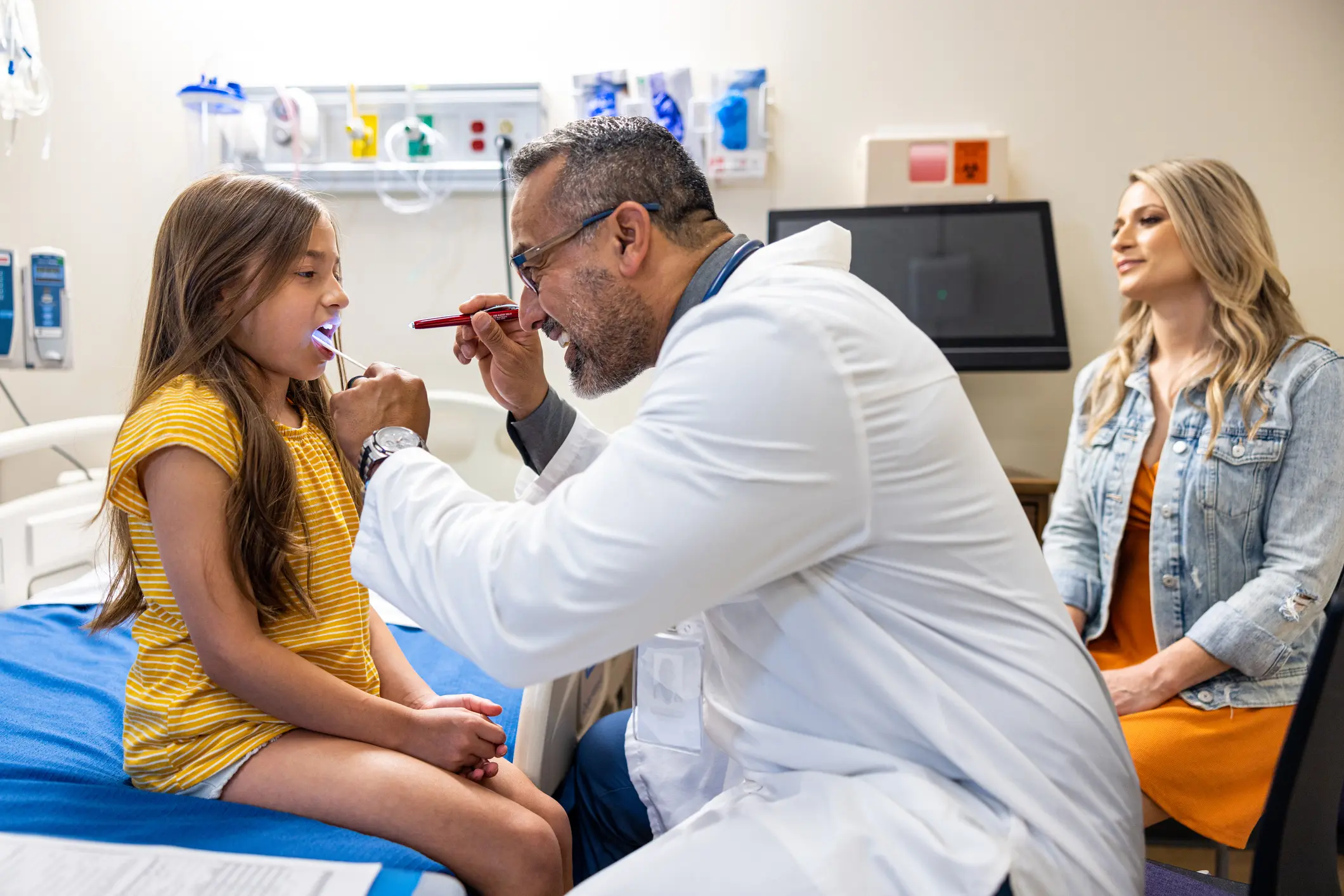 Ein Kinderarzt untersucht ein Mädchen auf einer Behandlungsliege, die Mutter sitzt beruhigend im Hintergrund – Routinebesuch beim Arzt