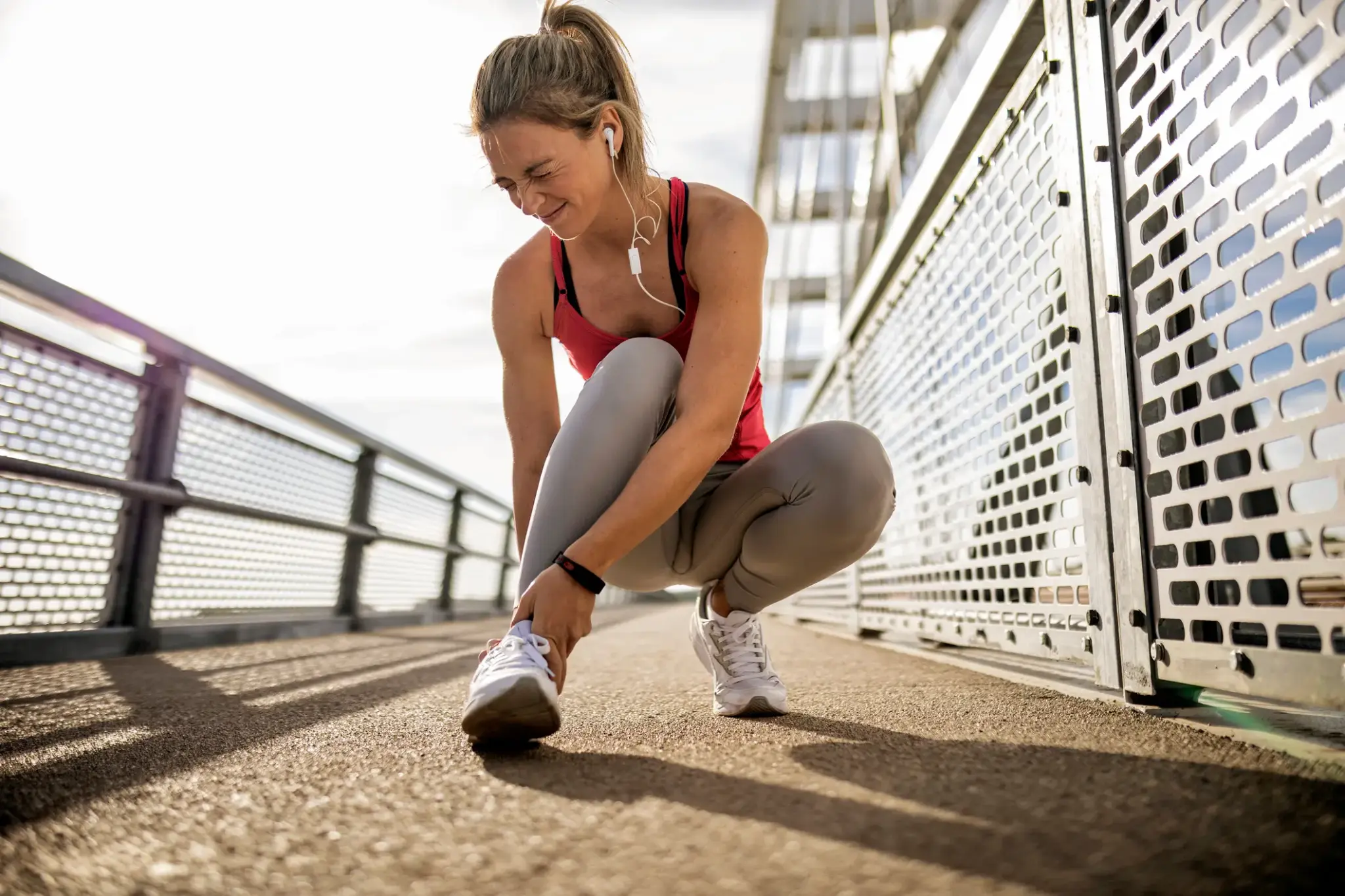 Junge Frau im Sportoutfit hält sich schmerzhaft den Knöchel nach einem Sturz beim Joggen – Sportunfall auf Brücke