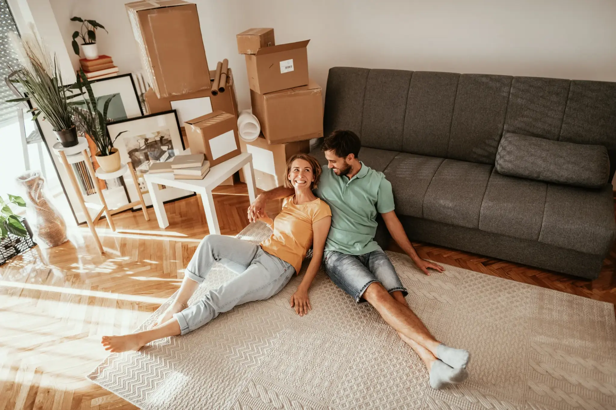 Junges Paar sitzt glücklich auf dem Teppich im Wohnzimmer zwischen Umzugskartons in der ersten eigenen Wohnung