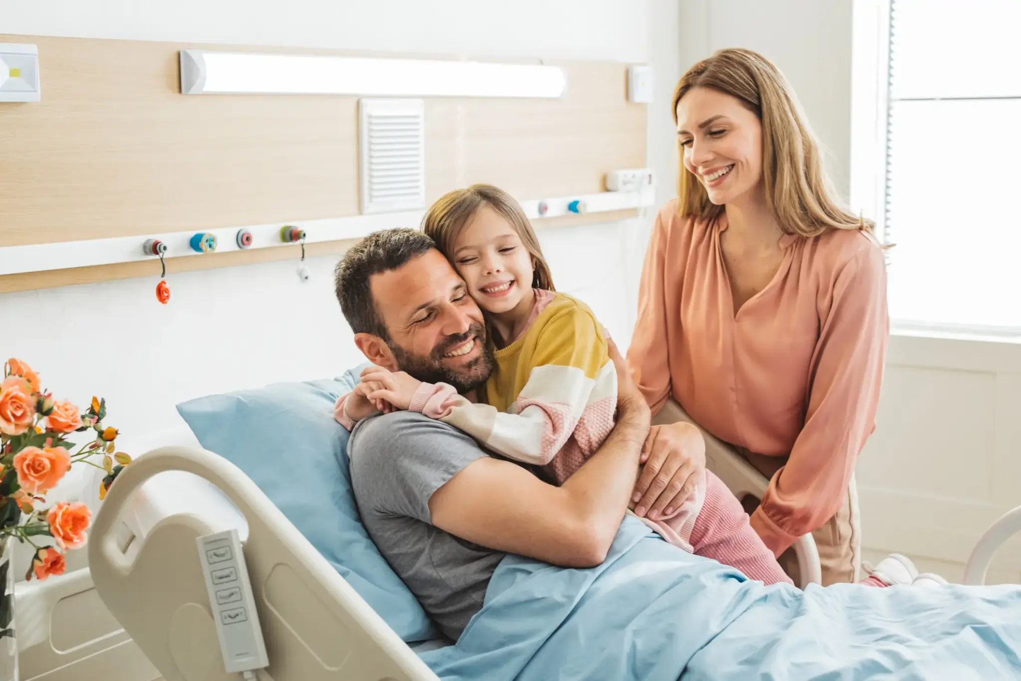 Mann liegt im Krankenhausbett und wird von seiner Tochter und Partnerin umarmt, alle lachen, helle Patientenzimmer-Atmosphäre
