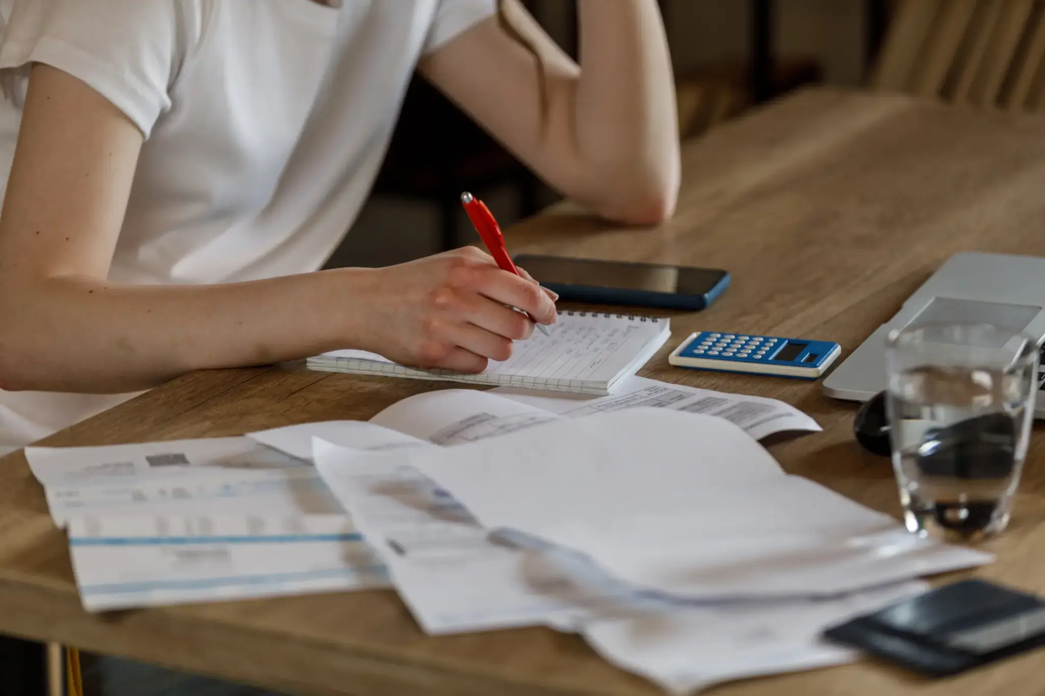 Person mit Block und Stift bei der Finanzplanung – auf dem Tisch liegen Unterlagen, Taschenrechner, Laptop und ein Glas Wasser.