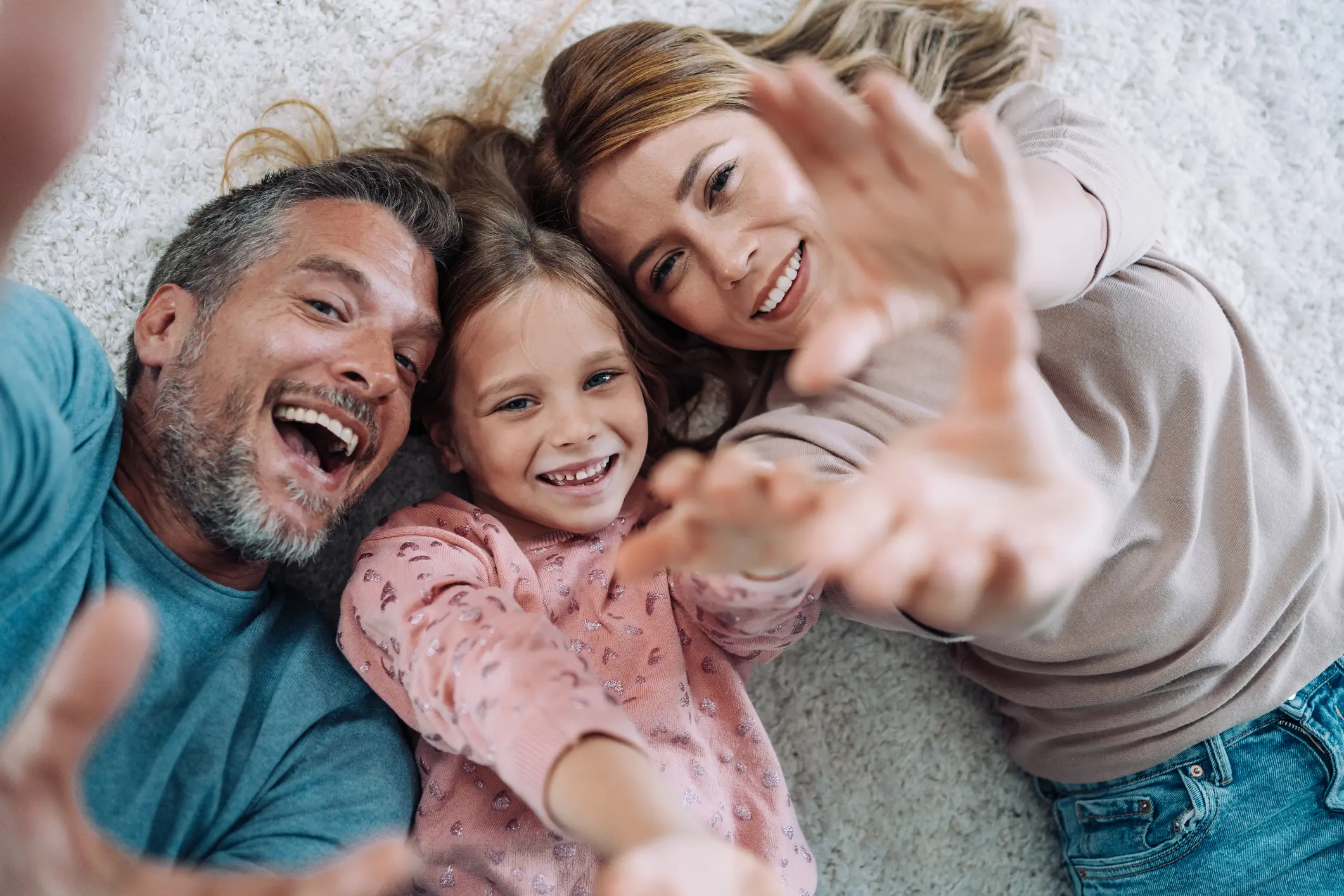Lächelnde Familie mit Mutter, Vater und Tochter liegt auf dem Teppich und streckt lachend die Hände in Richtung Kamera – Symbol für Geborgenheit.