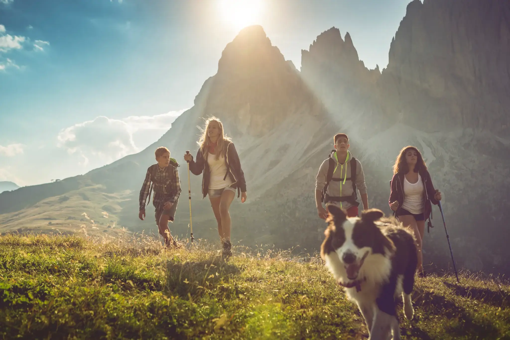Eine Wandergruppe mit Hund läuft bei Sonnenschein durch eine Berglandschaft, im Hintergrund ragen steile Felsen empor.
