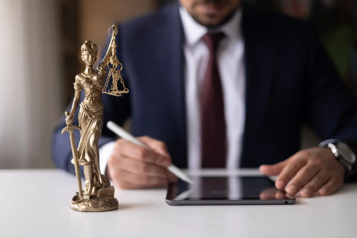 Justitia-Figur steht auf einem Schreibtisch, im Hintergrund arbeitet ein Mann im Anzug mit Tablet