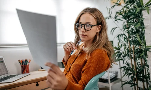 Junge Frau mit Brille sitzt am Schreibtisch, hält ein Dokument in der Hand und denkt nach, Laptop und Büroausstattung im Hintergrund