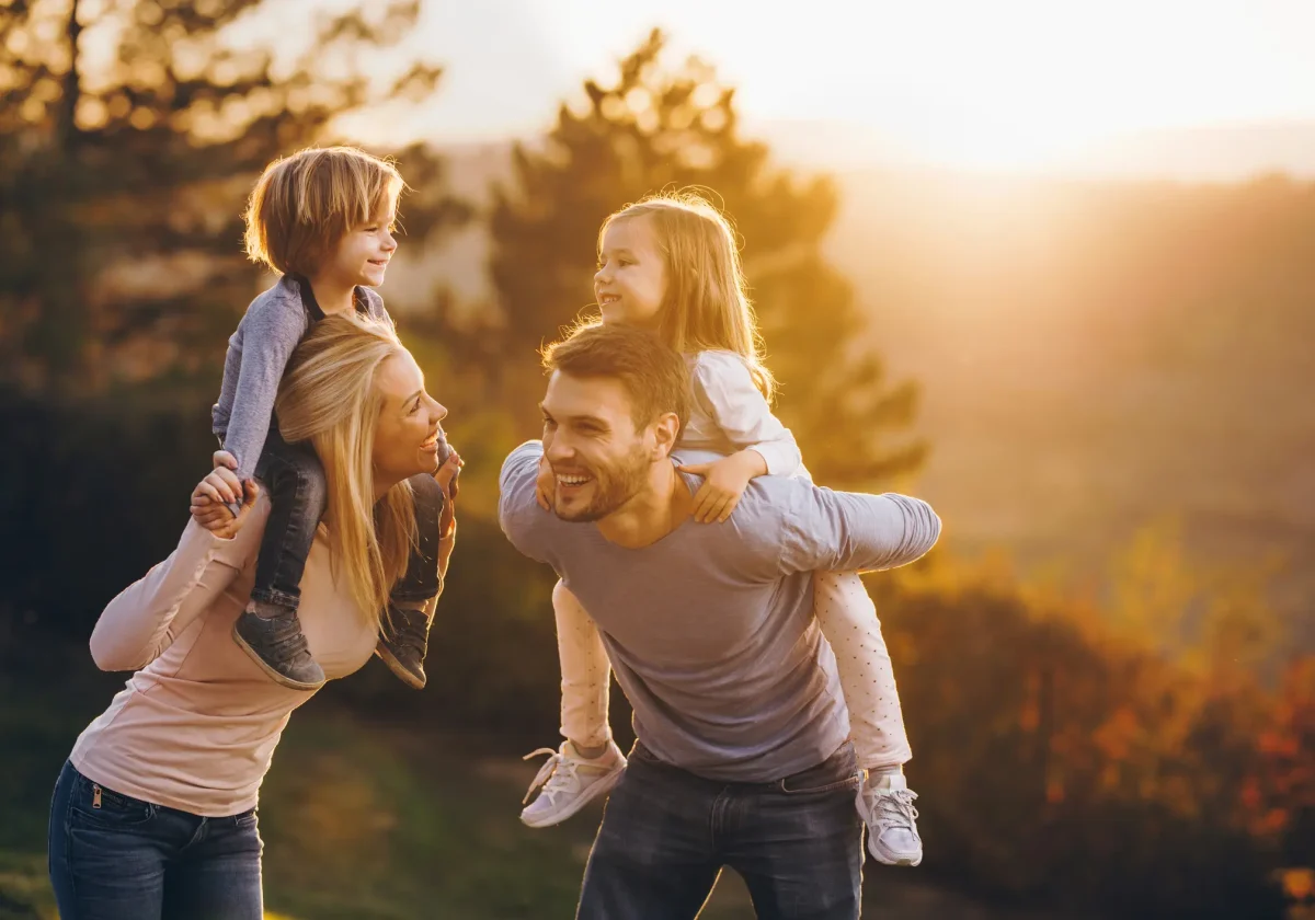BU-Versicherung Glückliche Familie spielt bei Sonnenuntergang im Freien – Vater trägt Tochter, Mutter trägt Sohn, Symbolbild für finanzielle Absicherung.