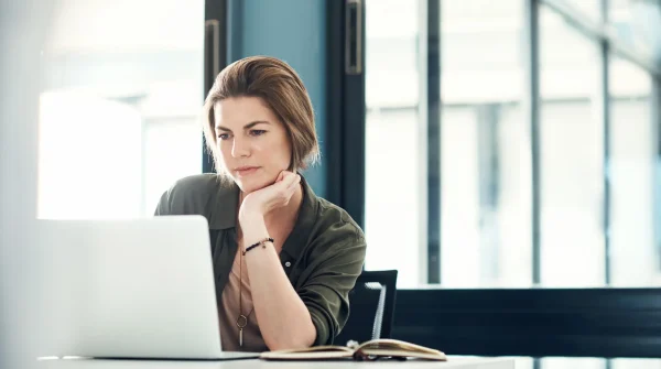 Frau sitzt am Schreibtisch und schaut nachdenklich auf einen Laptop, aufgeschlagenes Notizbuch vor ihr, moderne Büroumgebung
