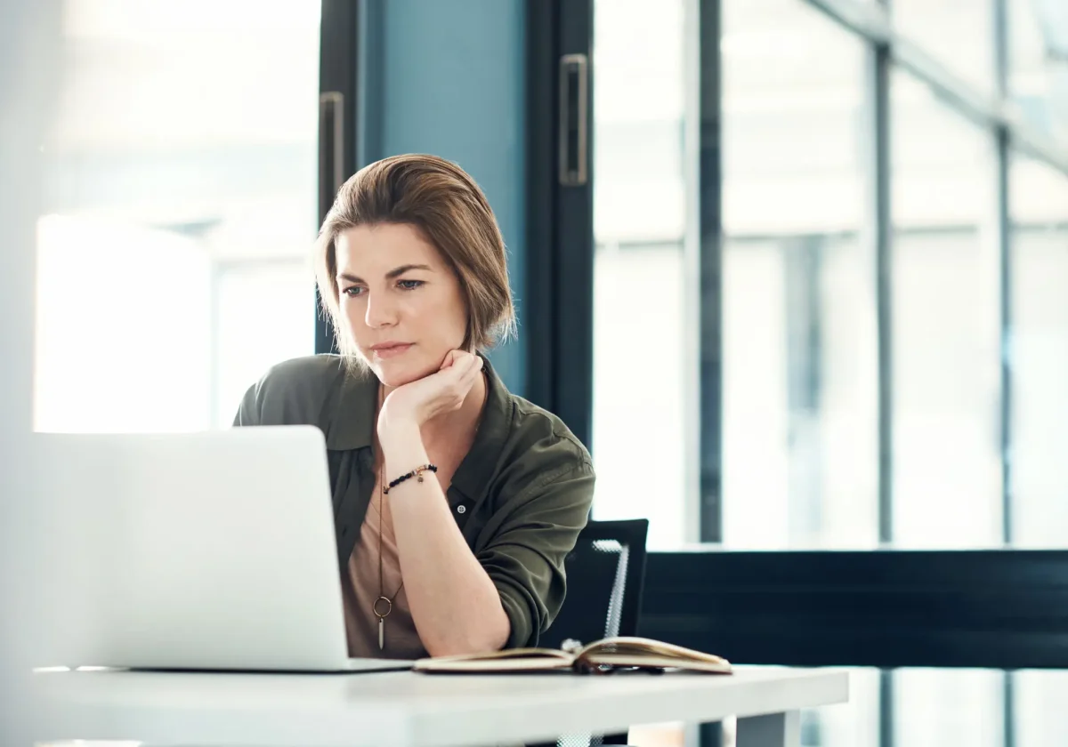 Frau sitzt am Schreibtisch und schaut nachdenklich auf einen Laptop, aufgeschlagenes Notizbuch vor ihr, moderne Büroumgebung