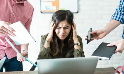 Gestresste Frau hält sich am Schreibtisch den Kopf, während ihr mehrere Aufgaben gleichzeitig zugeschoben werden