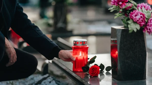 Hand stellt Grablicht neben rote Rose auf Grabstein – symbolischer Moment zur Erinnerung und Hinweis auf emotionale wie finanzielle Aspekte der Bestattung