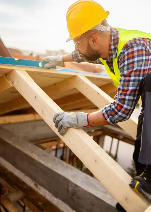 Dachdecker misst und richtet Holzbalken auf einem Neubau aus, mit Schutzkleidung, Helm und Wasserwaage bei sonnigem Wetter