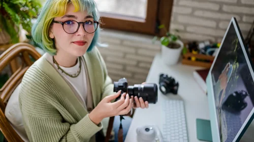 Junge Fotografin mit grünblonden Haaren sitzt an einem Schreibtisch und hält eine Kamera in der Hand, im Hintergrund ein Monitor mit Bildbearbeitung