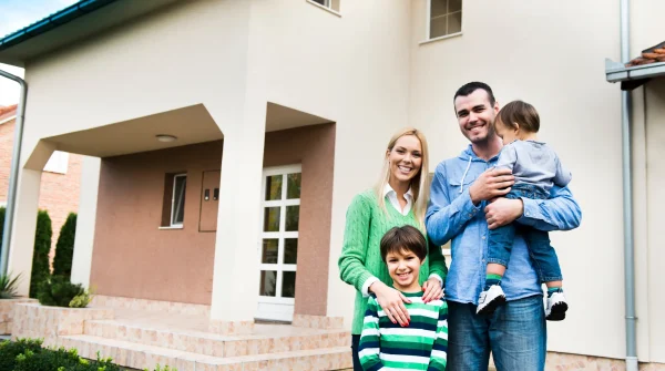 Familie mit zwei Kindern steht lächelnd vor dem eigenen Wohnhaus mit hell verputzter Fassade