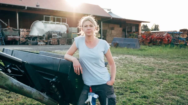 Landwirtin steht vor einem landwirtschaftlichen Gebäude mit Maschinen im Hof und lehnt an einem Traktor-Anbaugerät