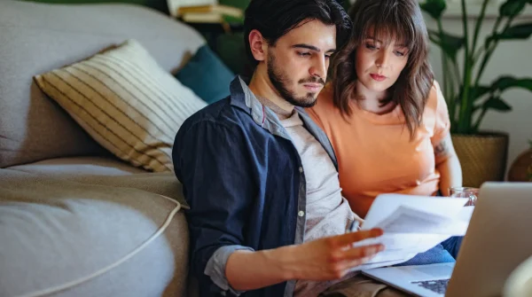 Junges Paar prüft Versicherungsunterlagen auf dem Sofa – Vergleich von Gebäudeversicherungen am Laptop.
