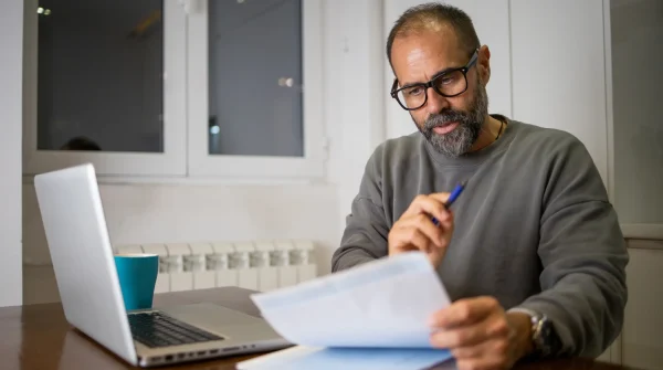 Mann mit Brille prüft Dokumente am Schreibtisch vor Laptop, im Hintergrund eine Kaffeetasse