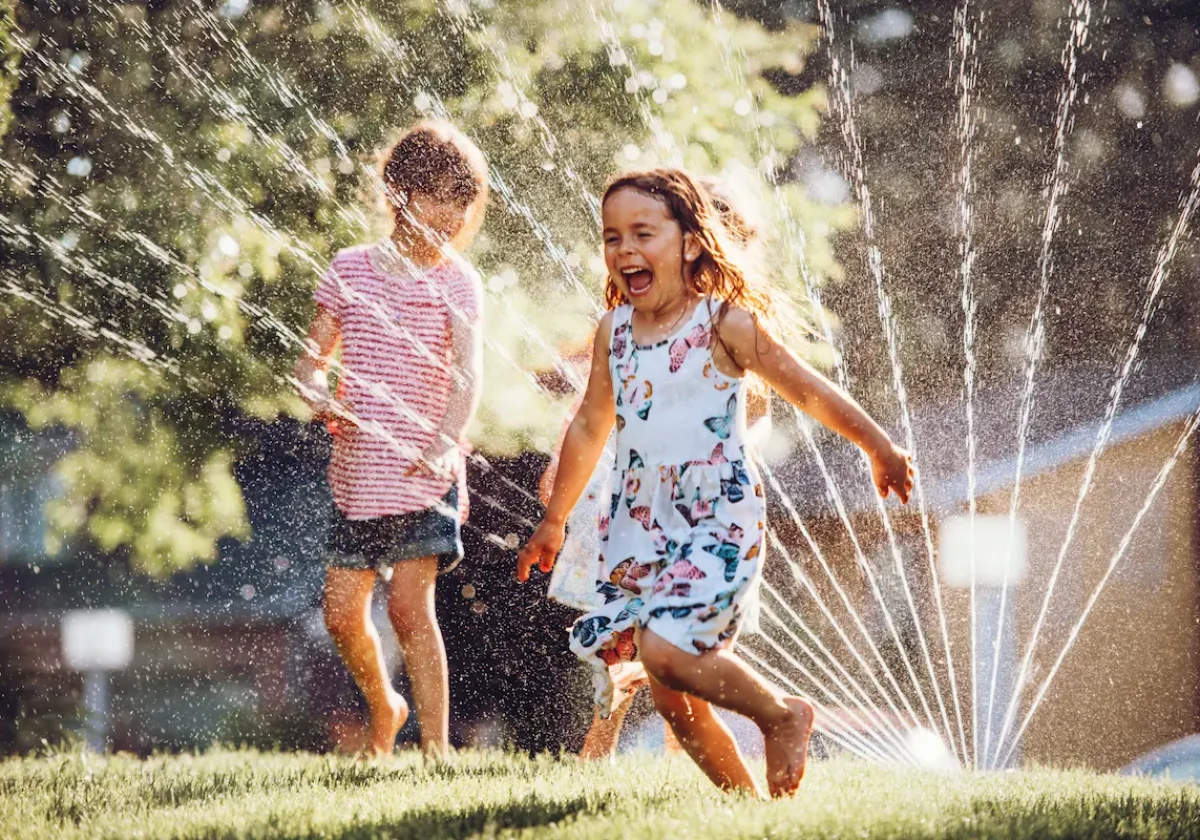 Zwei Kinder rennen lachend durch einen Rasensprenger, umgeben von Sonnenlicht und Wassertropfen