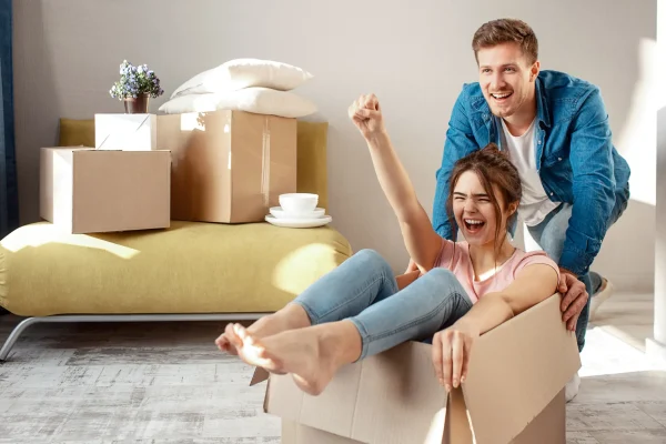 Lachendes Paar beim Umzug, Frau sitzt freudig in einem Umzugskarton, Kartons im Hintergrund
