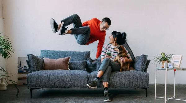 Paar mit Hund auf Sofa, spielerische Szene im Wohnzimmer – Symbolbild für Hausrat- und Haftpflichtversicherung im Alltag