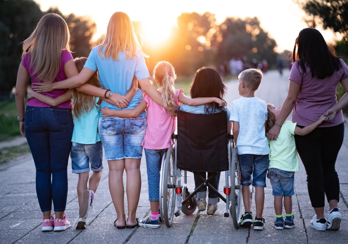 Kinderinvaliditaetsversicherung Kindergruppe von hinten, ein Kind im Rollstuhl wird liebevoll von den anderen umarmt – gemeinsamer Spaziergang bei Sonnenuntergang.