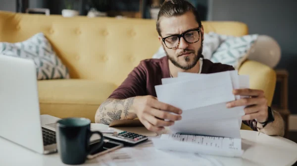Junger Mann mit Brille prüft Rechnungen am Wohnzimmertisch, neben Laptop und Taschenrechner – symbolisiert die Kostenfrage bei BU-Versicherungen