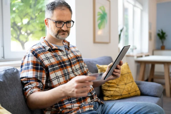 Mann mittleren Alters vergleicht Krankenkassen am Tablet und hält dabei eine Karte in der Hand – Symbolbild für Krankenkassenvergleich online
