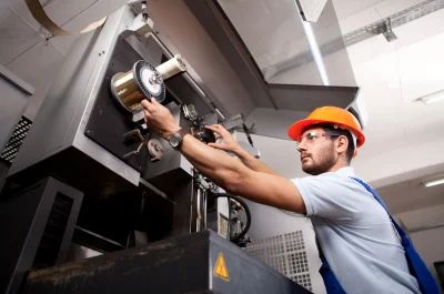 Techniker mit Schutzhelm prüft in der Industriehalle eine laufende Produktionsmaschine auf Funktion und Sicherheit