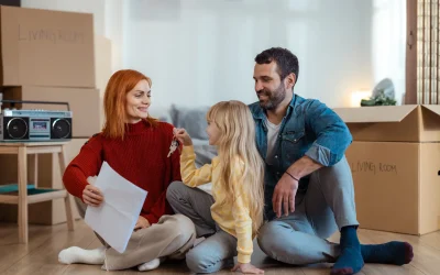 Junge Familie sitzt beim Einzug zwischen Umzugskartons – Kind überreicht symbolisch die Wohnungsschlüssel an die Mutter