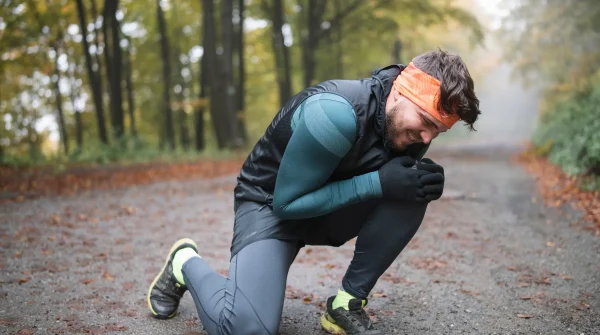 Mann kniet verletzt im Herbstwald auf dem Weg und hält sich schmerzverzerrt das Knie – Joggingkleidung und Stirnband