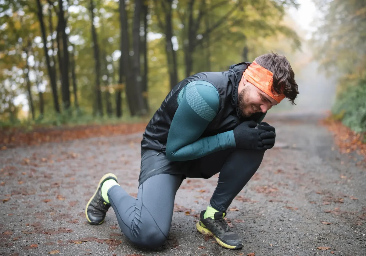 Mitwirkungsanteil Mann kniet verletzt im Herbstwald auf dem Weg und hält sich schmerzverzerrt das Knie – Joggingkleidung und Stirnband