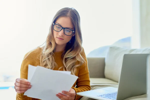 Junge Frau mit Brille prüft konzentriert Dokumente vor dem Laptop – Entscheidung zur Anpassung einer Berufsunfähigkeitsversicherung.