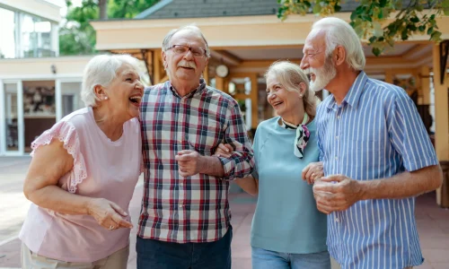 Vier ältere Menschen lachen gemeinsam im Freien und gehen Arm in Arm, gepflegte Umgebung im Hintergrund