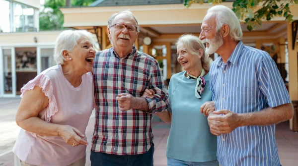 Vier ältere Menschen lachen gemeinsam im Freien und gehen Arm in Arm, gepflegte Umgebung im Hintergrund