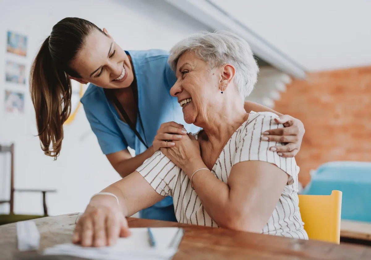 Junge Pflegerin umarmt lächelnde Seniorin an einem Tisch, warmherzige Pflegesituation in häuslicher Umgebung