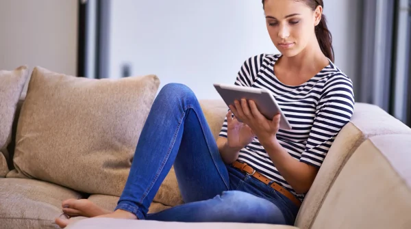 Junge Frau sitzt entspannt auf dem Sofa und nutzt ein Tablet – Symbolbild für digitale Information und persönliche Absicherung.