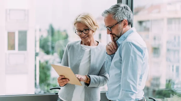 Älteres Geschäftspaar bespricht gemeinsam Informationen auf einem Tablet vor Bürofenster
