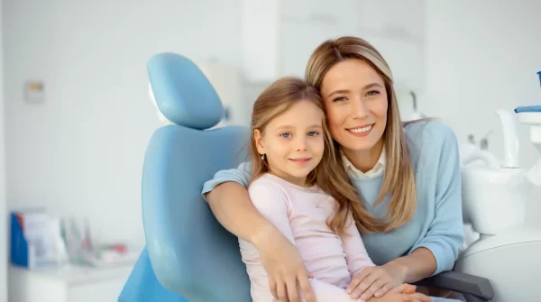 Mutter mit Tochter beim Zahnarzt – gemeinsam lächeln sie im Behandlungszimmer und warten auf die Beratung zur Zahnzusatzversicherung.